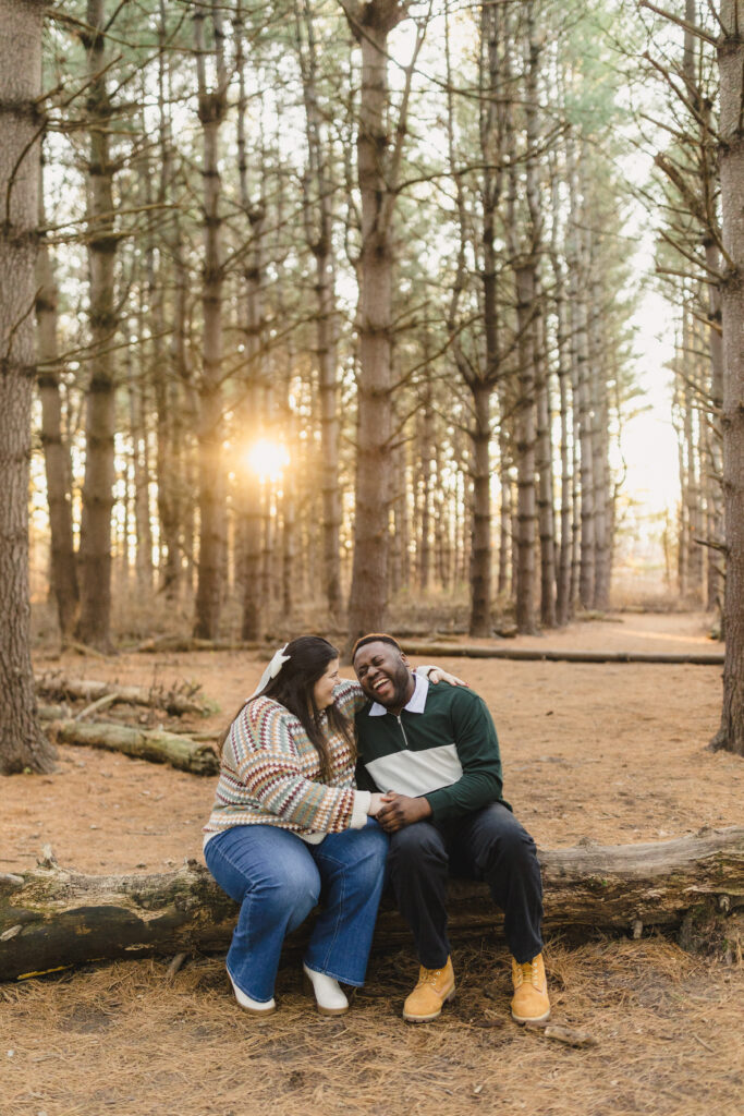 a couple sitting on a tree trunk in the woods laughing with one another during their engagement session 
