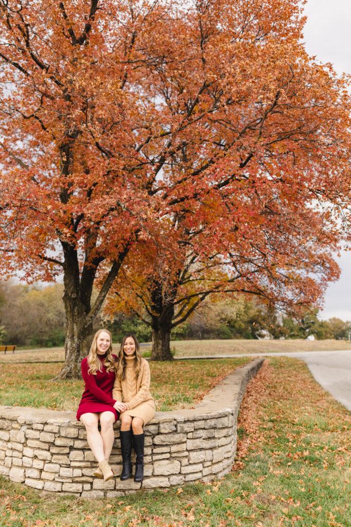 two women sitting and smiling on a stone wall during their kansas city fall engagement session 