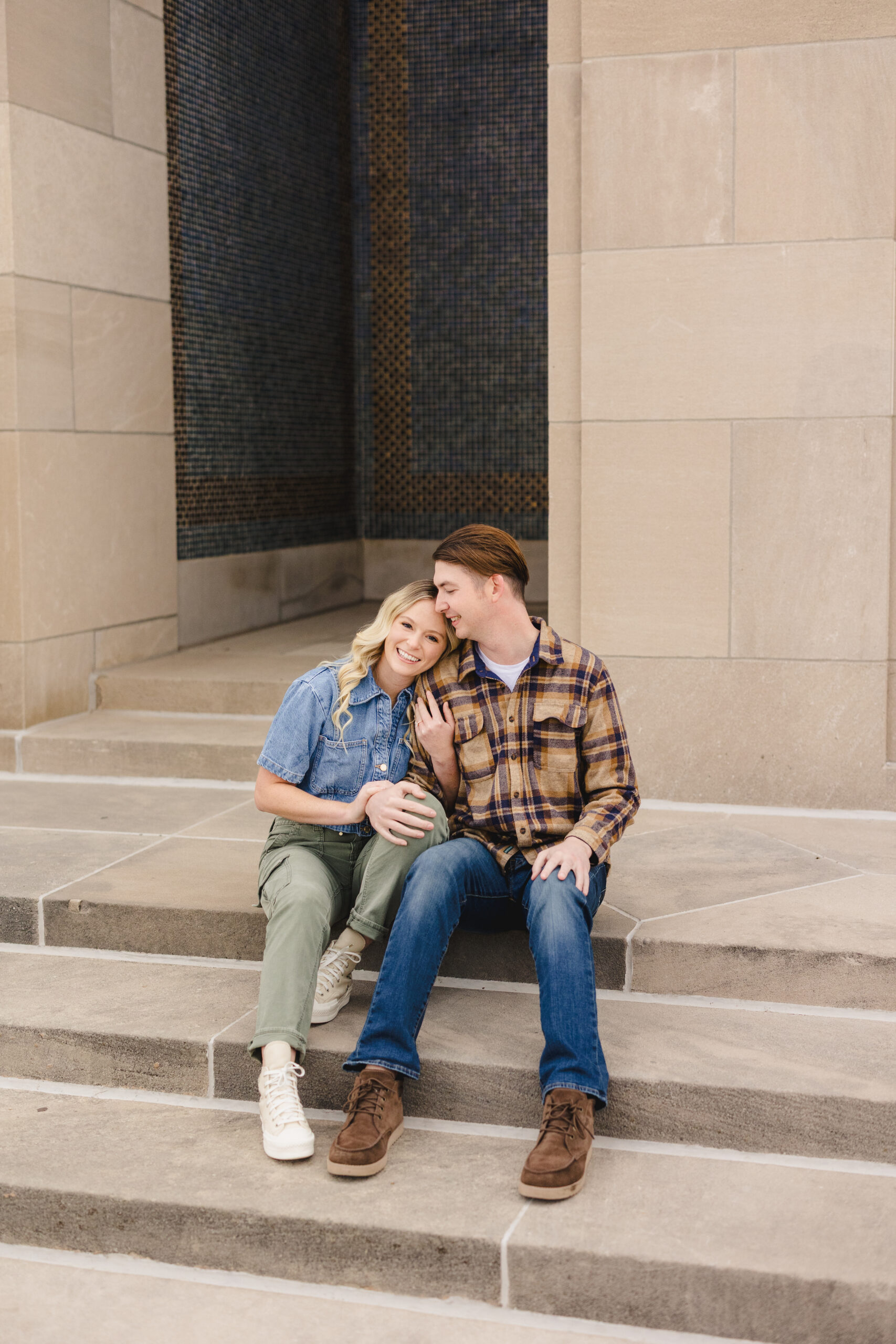 a couple sitting on steps together taking fall engagement photos the woman has her hands wrapped around her partner's arm 