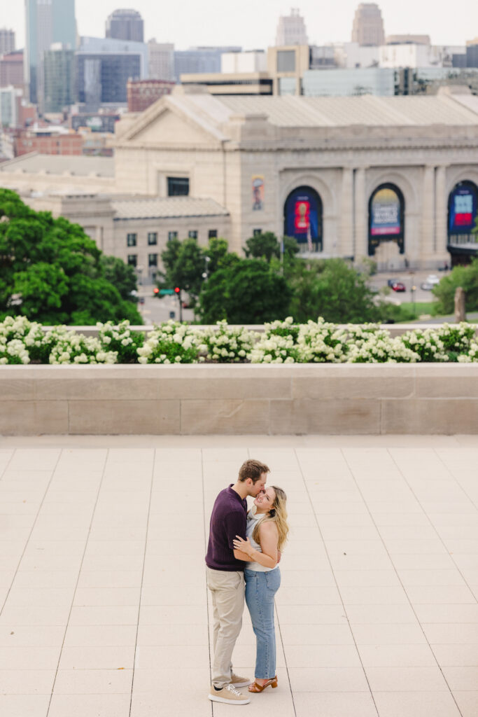 a man kissing his partner on the cheek with the kansas city skyline behind them 