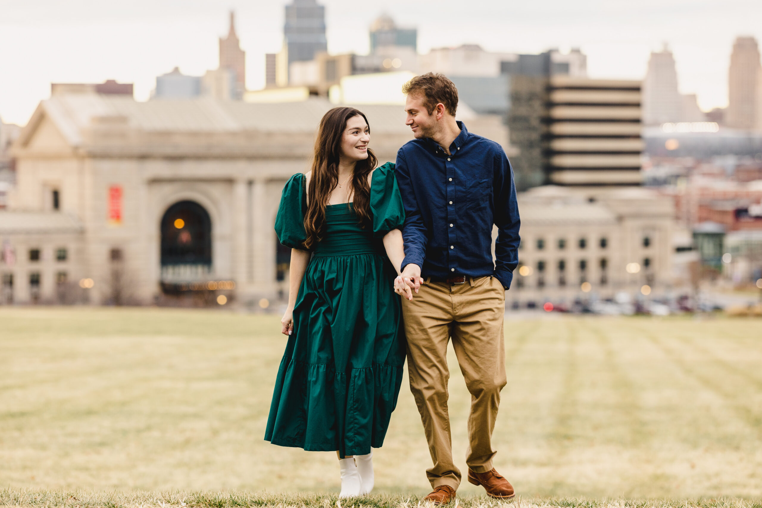 a couple walking hand in hand on a lawn with the kansas city skyline behind them 
