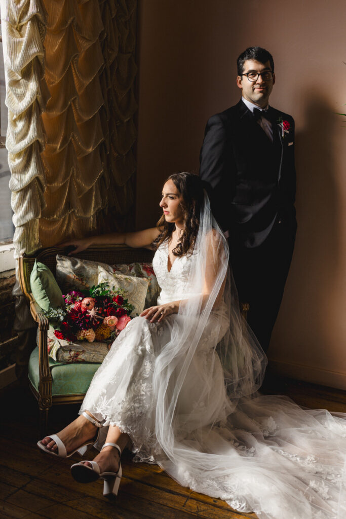 A person in a wedding dress sitting in a chair looking out a window with their partner standing next to them 
