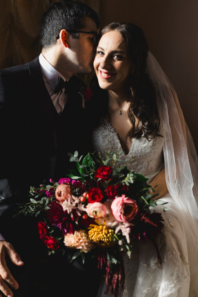 A person kissing their new spouse as they smile and hold a bouquet of flowers 
