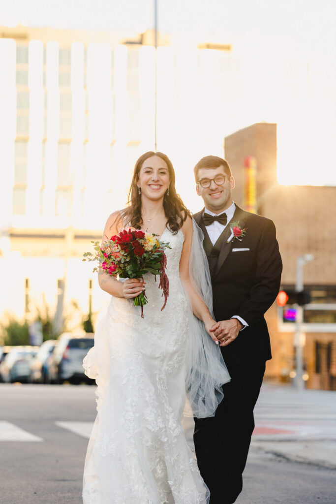 Newlyweds holding hands and smiling as they walk down a city street 

