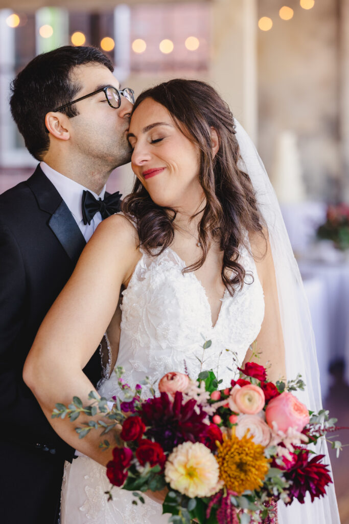a couple taking wedding portraits inside of the bauer in kansas city the groom is kissing his bride on the side of her head 