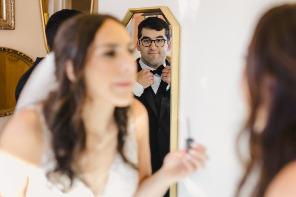A person putting on lipgloss with their partner's reflection in a mirror behind them adjusting their bow tie 
