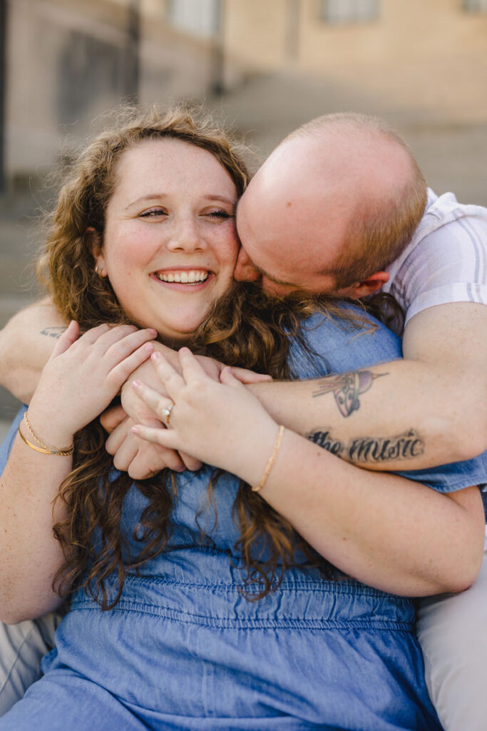 A person sitting behind their partner with their hands over their shoulders as they kiss their cheeks
