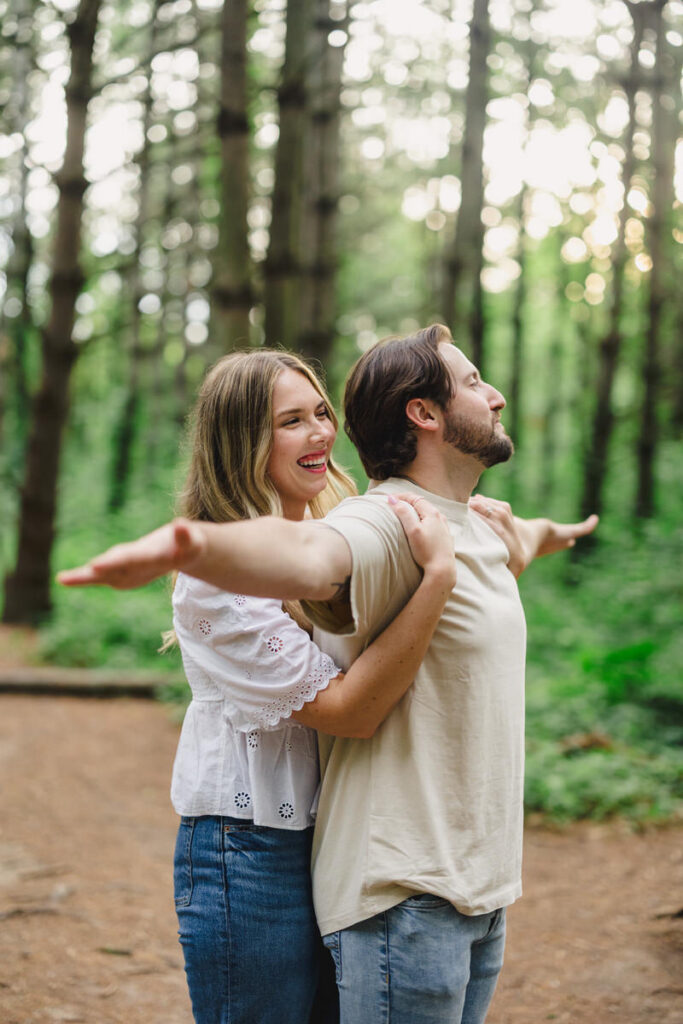 A couple standing in the titanic pose laughing while in the woods