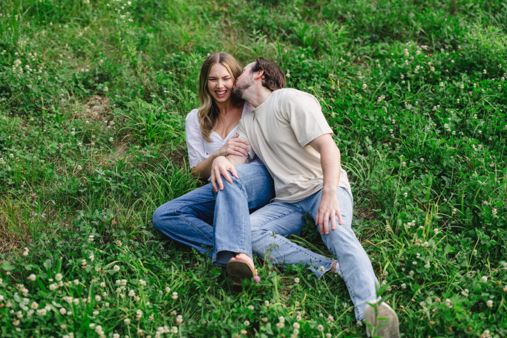 A couple sitting in the grass as one leans back to kiss the other's cheek