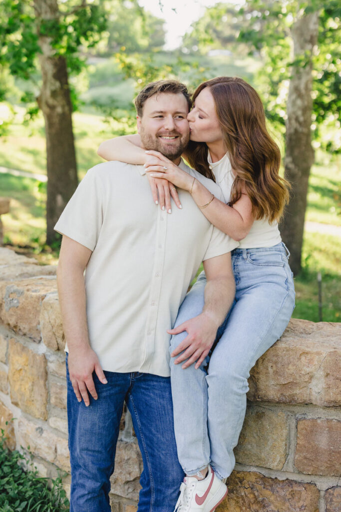 A person sitting on a small stone wall with their arms around their partner as they kiss their cheek