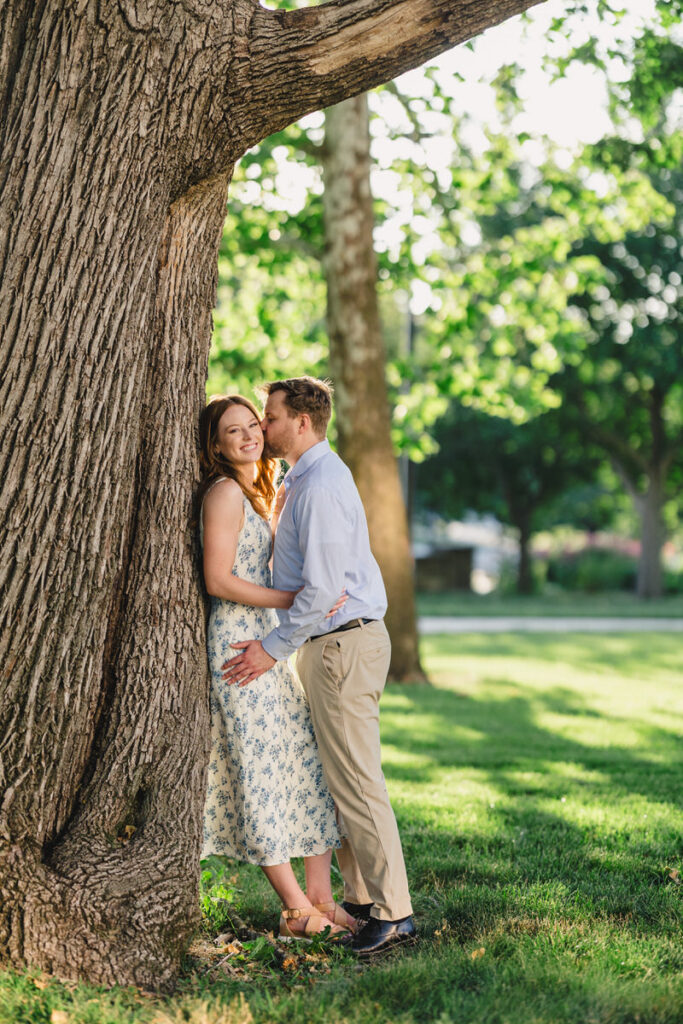 A couple leaning against a tree as one kisses the other's cheek