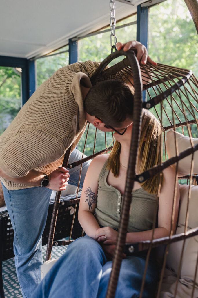 A person sitting in a hanging chair as their partner leans over them and kisses them