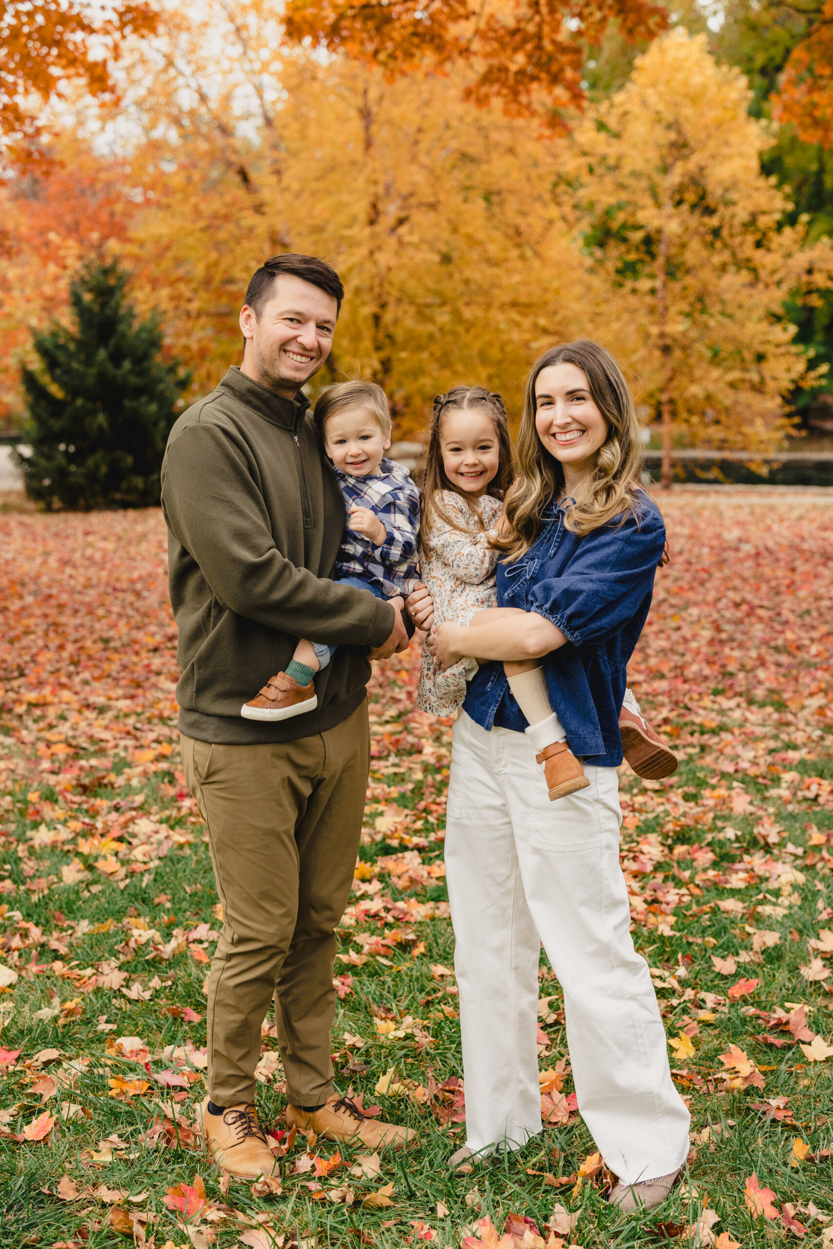 a mother and father in loose park during the fall holding their two children and taking fall family photos 