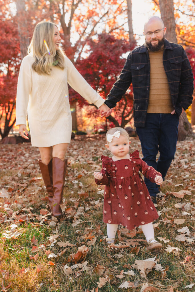 Parents holding hands and walking behind their toddler in a park 