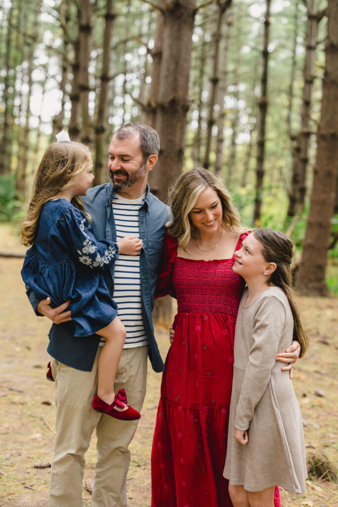 Parents standing in a wooded area each smiling at a child next to them 