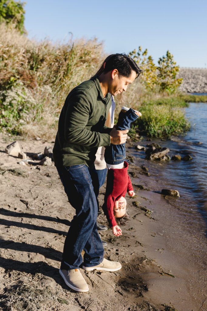 A parent holding their toddler upside down at the edge of a beach as they smile 