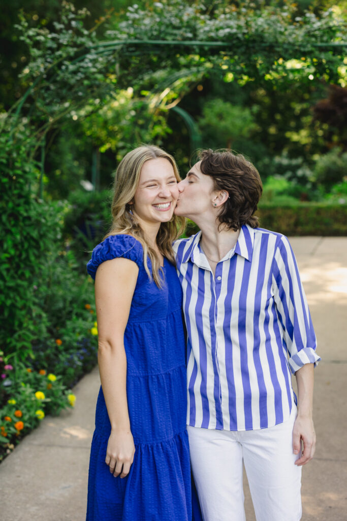 A couple with their arms around each other as one kisses the other's cheek 