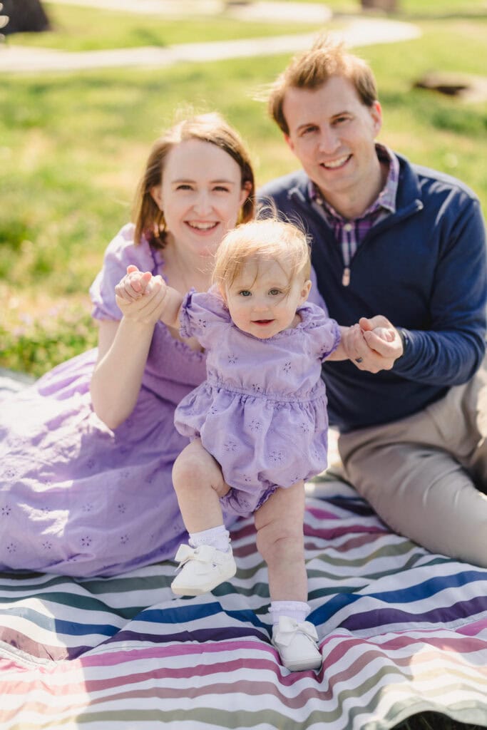 Parents sitting on a blanket in a park each holding their toddler's hand in front of them 