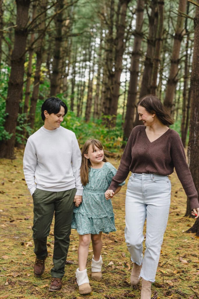 A child standing in between their parents as they all walk hand in hand in a wooded area 
