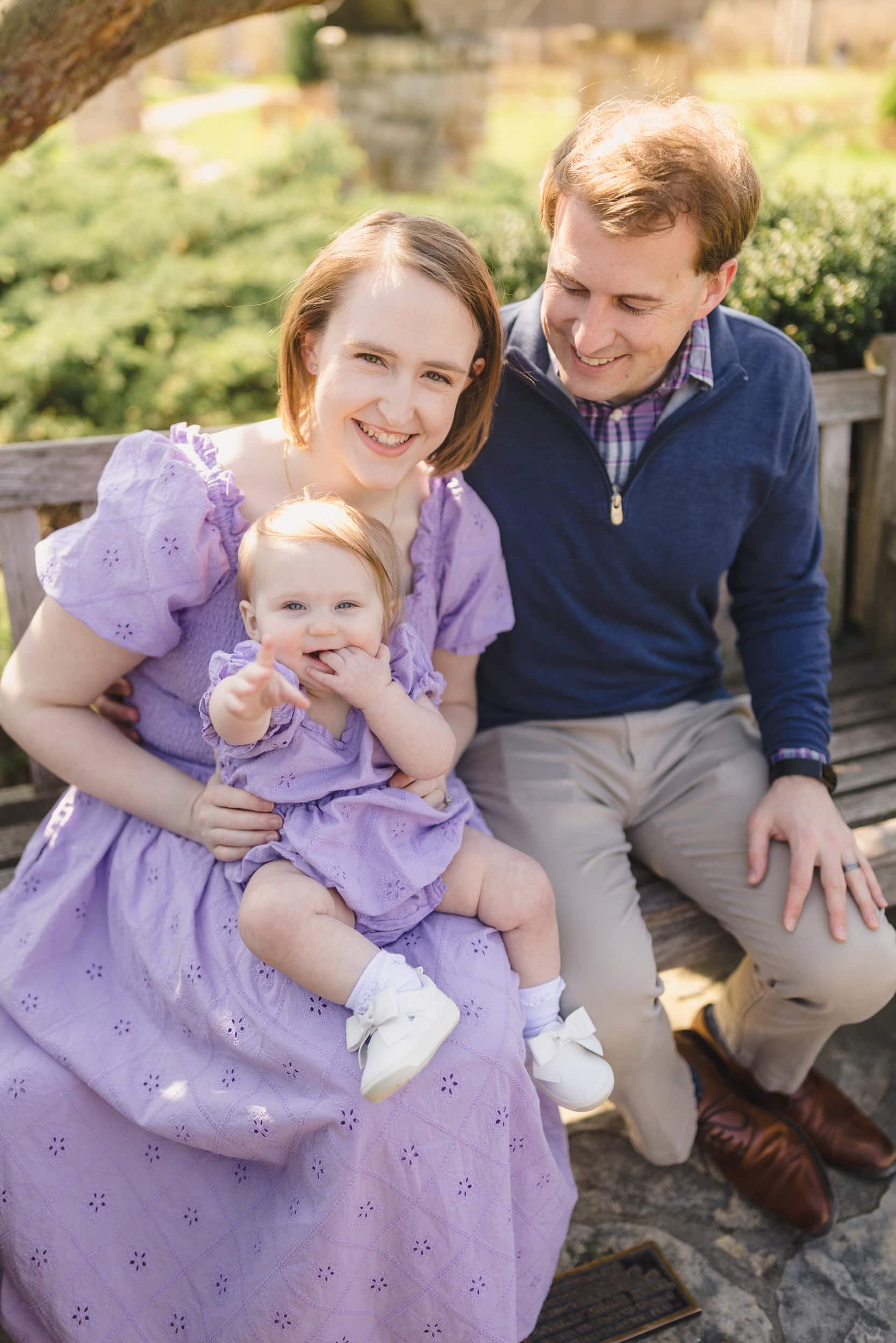A toddler sitting on their parents laugh as they laugh and reach one hand out
