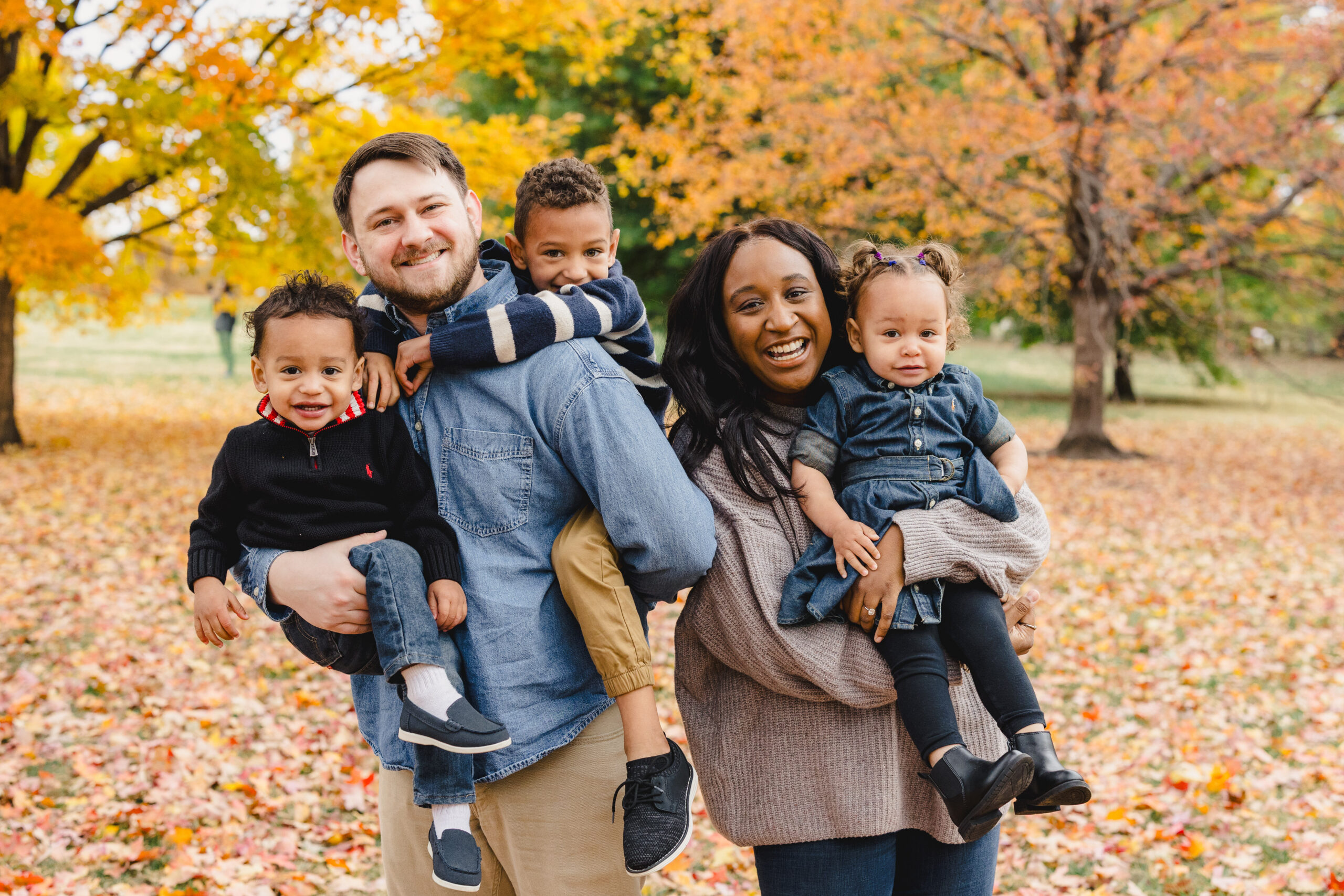 a mother and father holding their 3 children in loose park during the fall